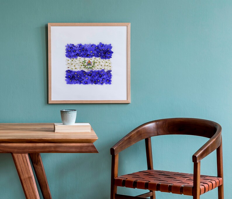 May include: A framed print of a flag made of purple and white flowers. The flag is displayed on a light blue wall above a wooden table and chair.
