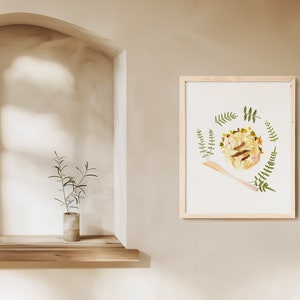 May include: A framed print of a pressed flower arrangement in a circle with a wooden spoon in the bottom right corner. The flowers are yellow, white, and brown, and the leaves are green.