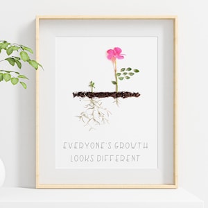 Op de afbeelding: Een ingelijst print met een witte achtergrond en een roze bloem met groene bladeren. De bloem groeit uit de grond en heeft wortels. De tekst "Everyone's growth looks different" is onder de bloem gedrukt.