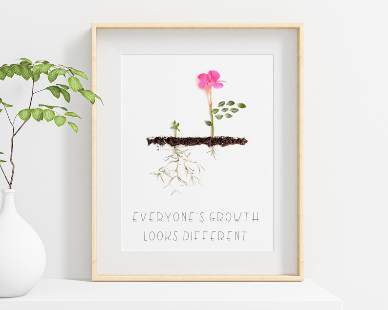 Op de afbeelding: Een ingelijst print met een witte achtergrond en een roze bloem met groene bladeren. De bloem groeit uit de grond en heeft wortels. De tekst "Everyone's growth looks different" is onder de bloem gedrukt.