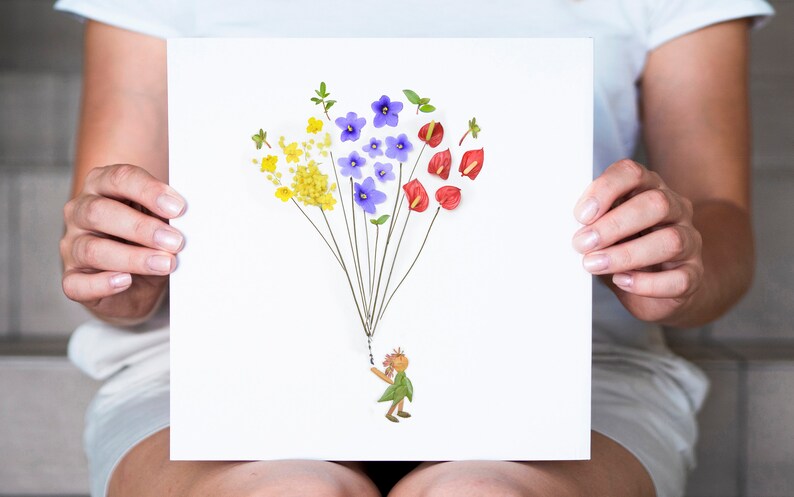 May include: A white card with a flower arrangement depicting a person holding a bunch of yellow, purple, and red flowers. The flowers are arranged to look like a hot air balloon.