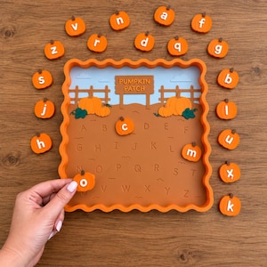 May include: A square orange tray with a scalloped edge featuring a pumpkin patch illustration and the text PUMPKIN PATCH. The tray contains an alphabet grid. Twenty-six individual orange pumpkin-shaped tokens, each marked with a lowercase letter, are arranged around the tray, with one token being held.