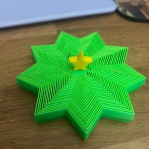 May include: A layered, star-shaped object in shades of green and yellow, with a small yellow star in the center. The object has a three-dimensional appearance, with multiple layers creating a textured effect. The design is geometric and modern.