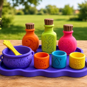May include: A set of colorful silicone potion-making toys including a purple cauldron with a yellow stirring spoon, three bottles with brown corks in orange, green, and pink, and three small cups in orange, blue, and yellow on a purple tray.