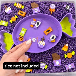 May include: A purple candy-shaped tray containing various Halloween-themed silicone charms including skulls, bats, pumpkins, chocolate bars, and lollipops. The tray is surrounded by purple rice. The text "rice not included" is visible.