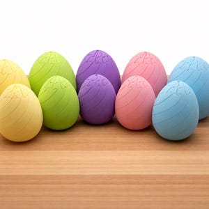 May include: A collection of colorful, egg-shaped objects in yellow, green, purple, pink, and blue. Each egg has a textured surface with curved lines and a decorative top. The eggs are arranged on a wooden surface.