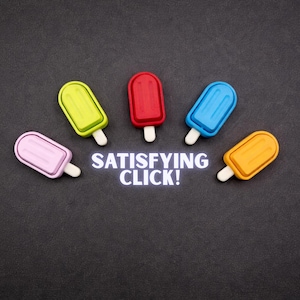 May include: An overhead shot features five colorful popsicle-shaped erasers arranged around the text "SATISFYING CLICK!". The erasers are pink, green, red, blue, and yellow, each with a white stick, set against a dark gray background.