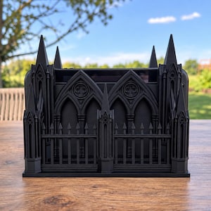 May include: A matte black desk organizer shaped like a gothic cathedral facade, featuring pointed arches, intricate window tracery, and a spiked fence perimeter. The structure has a rectangular open top compartment for holding pens or stationery items.