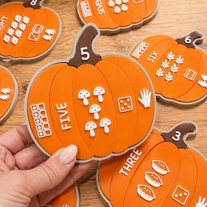 May include: A set of orange pumpkin-shaped educational counting cards. Each card features a large numeral on the stem, the written number word, a ten-frame grid, a group of small white icons representing the quantity, a die face, and a hand showing fingers.