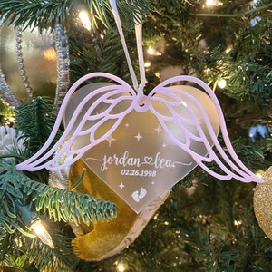 May include: A pink and white heart-shaped Christmas ornament with angel wings. The ornament is personalized with the names "Jordan & Lea" and the date "02.26.1998".