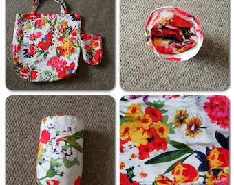May include: A floral print tote bag with a smaller matching pouch. The bag has a white background with red, yellow, and green flowers. The bag is folded and laid flat on a light brown surface.