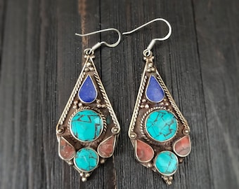May include: A pair of silver-toned earrings with turquoise, blue, and red stones. The earrings have a teardrop shape with a triangular top, a central circular turquoise stone, and smaller turquoise and red stones at the bottom.