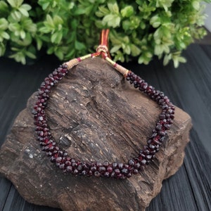 May include: A red garnet gemstone necklace with a gold and red cord closure. The necklace is displayed on a dark brown rock.