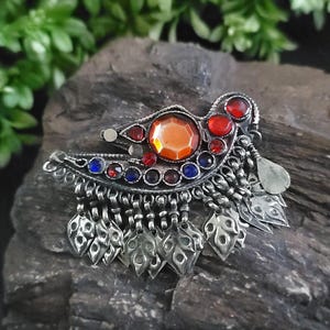 Puede incluir: Broche plateado con una piedra central naranja, rodeada de gemas rojas y azules. El broche presenta colgantes plateados en forma de hoja y está sobre un fondo oscuro y texturizado. La joya tiene un diseño vintage ornamentado.