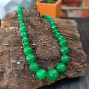 May include: A green jade bead necklace with round beads. The necklace is displayed on a piece of wood.
