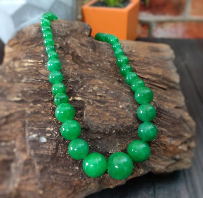 May include: A green jade bead necklace with round beads. The necklace is displayed on a piece of wood.
