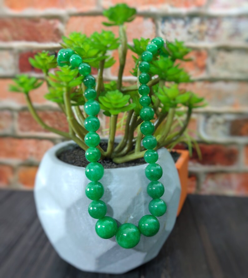 May include: A green jade bead necklace with round beads. The necklace is draped over a white planter with a green succulent plant.