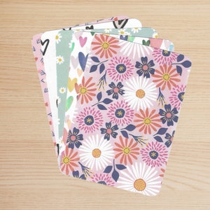 Può includere: Un set di sei carte decorative con una varietà di motivi floreali. Le carte sono rosa, verdi e bianche con una varietà di disegni floreali, tra cui margherite, cuori e foglie.