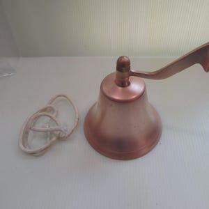 May include: A copper-colored bell with a handle and a white rope. The bell has a rounded base and a handle that curves upward. The rope is knotted. The bell is likely used for signaling or decoration.