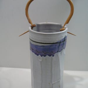 May include: A handmade ceramic vase with a light blue and purple painted design. The vase has a textured surface and a woven wooden handle. The handle is attached to the vase with leather straps. The vase is cylindrical in shape.