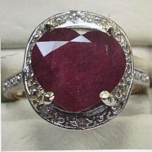Peut inclure: Gros plan sur une bague ornée d'une grosse pierre précieuse rouge foncé en forme de cœur. La pierre précieuse est entourée d'un halo de petites pierres claires serties dans un anneau en métal argenté. La bague a un anneau doré.
