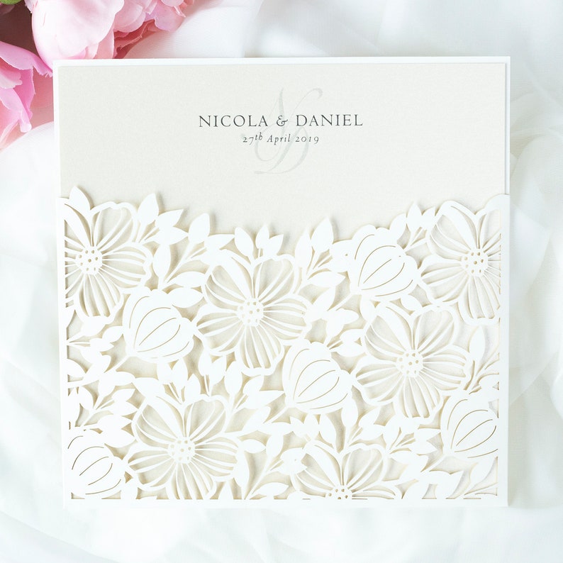 May include: Cream-coloured wedding invitation with a laser-cut floral design. The invitation features the names "Nicola & Daniel" and the date "27th April 2019". The intricate floral pattern adds a delicate touch to the design.