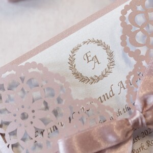 May include: A pink and white wedding invitation with a delicate lace design. The invitation features the initials "EA" in a wreath and the text "Evelyn & Alex" and "August 2022 Park Rd."