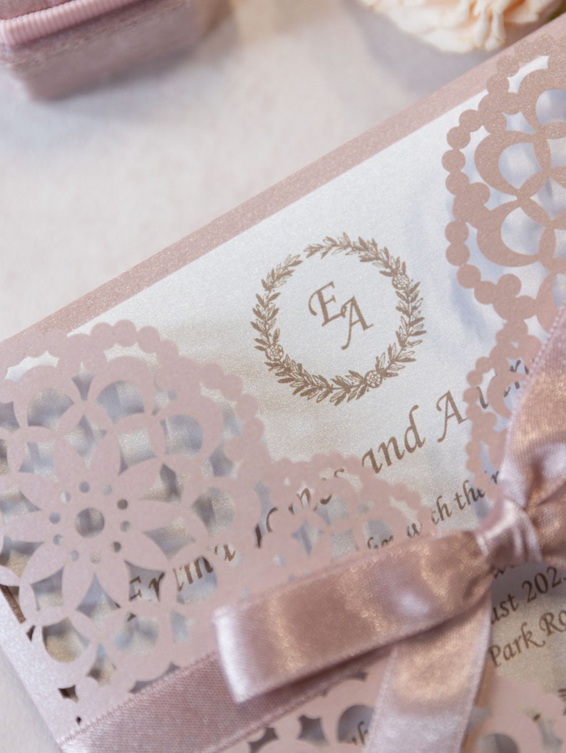 May include: A pink and white wedding invitation with a delicate lace design. The invitation features the initials "EA" in a wreath and the text "Evelyn & Alex" and "August 2022 Park Rd."