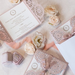 May include: A set of wedding invitations with a delicate lace design in a blush pink color. The invitations are folded and tied with a satin ribbon. The text on the invitation reads "Emma James and Andrew Sewell".