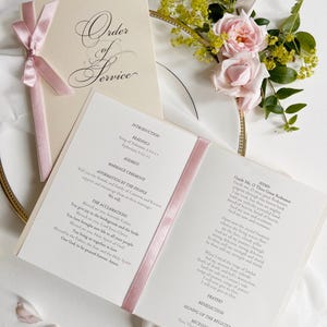 May include: A cream-colored wedding order of service booklet with a pink satin ribbon tie. The cover features the text Order of Service in elegant script. The open interior pages display printed ceremony details, readings, and hymn lyrics.