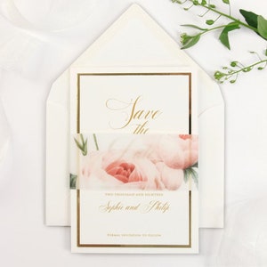 Op de afbeelding: Save the Date kaart met een bloemenontwerp. De kaart is wit met gouden letters en een roze bloemenlint. De tekst luidt "Save the Date" en "Twee Duizend Achttien Sophie en Philip Formele uitnodiging volgt."