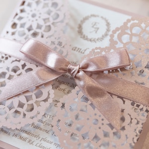 May include: A blush pink wedding invitation with a delicate lace design and a matching satin ribbon tied in a bow. The invitation features the couple's initials in a wreath and the text "RSVP" and "alternatively please reply".