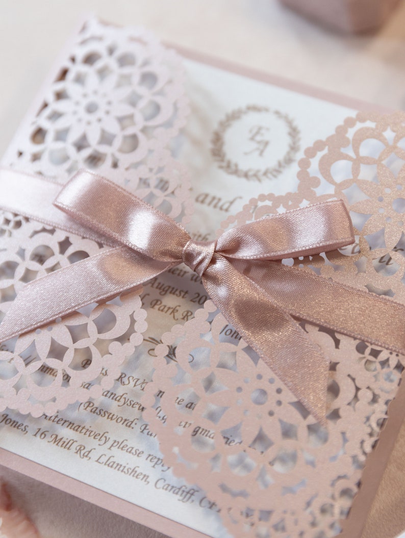 May include: A blush pink wedding invitation with a delicate lace design and a matching satin ribbon tied in a bow. The invitation features the couple's initials in a wreath and the text "RSVP" and "alternatively please reply".