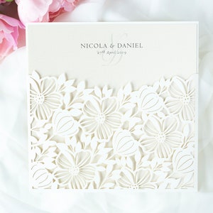 May include: Ivory wedding invitation with a laser-cut floral design. The invitation features the names "Nicola & Daniel" and the date "27th April 2019". The invitation is set against a white fabric background with pink flowers.