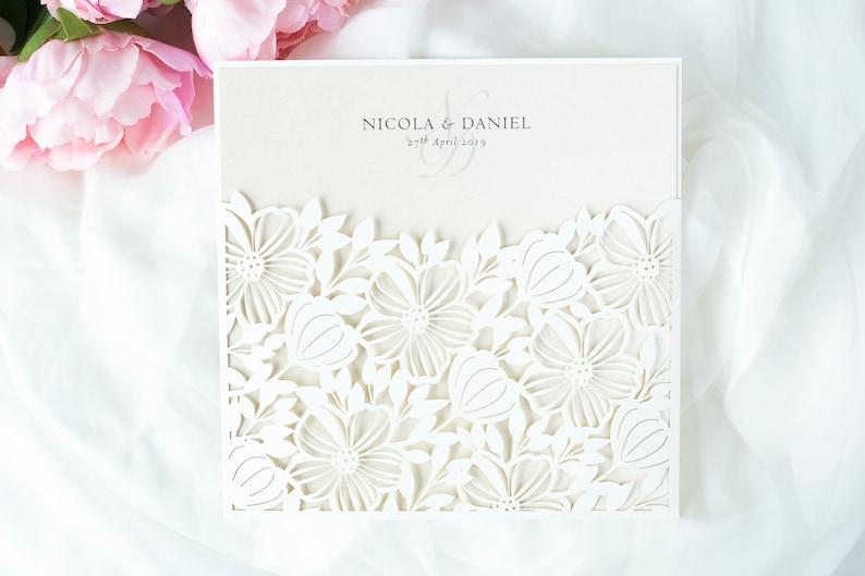 May include: Ivory wedding invitation with a laser-cut floral design. The invitation features the names "Nicola & Daniel" and the date "27th April 2019". The invitation is set against a white fabric background with pink flowers.