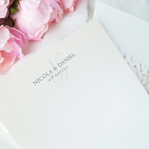 May include: Ivory wedding invitation with the names "Nicola & Daniel" and the date "27th April 2019" printed in grey. The invitation is next to a white envelope with a floral laser-cut design and pink peonies.