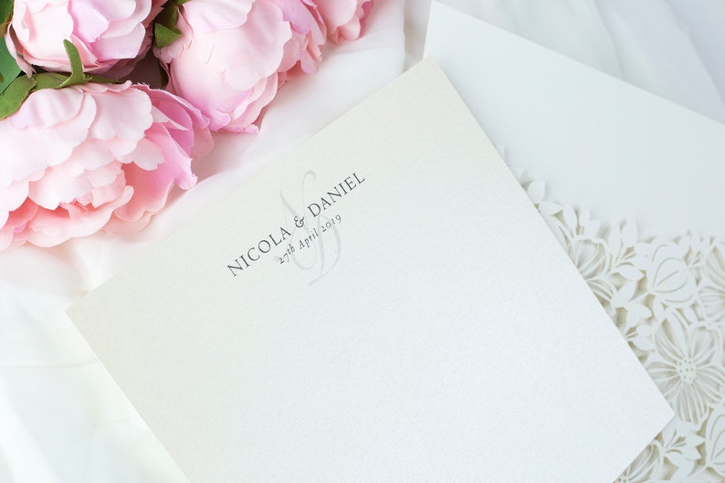 May include: Ivory wedding invitation with the names "Nicola & Daniel" and the date "27th April 2019" printed in grey. The invitation is next to a white envelope with a floral laser-cut design and pink peonies.