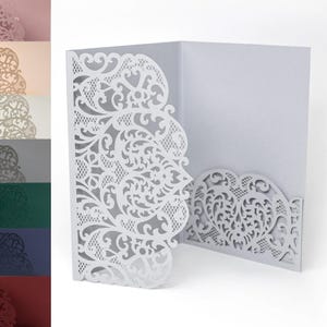 May include: A white laser-cut invitation card with intricate floral and scrollwork designs. The card is open, revealing a blank interior for text. The design features a decorative border and a heart-shaped pocket.