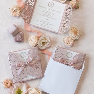 May include: A blush pink wedding invitation with a lace overlay and a white envelope. The invitation features the names "Emma James and Andrew Sewell" and the date "29th August 2021".