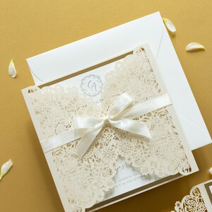 May include: A white wedding invitation with a delicate lace design and a white satin ribbon tied in a bow. The invitation features the initials "GA" in a wreath.