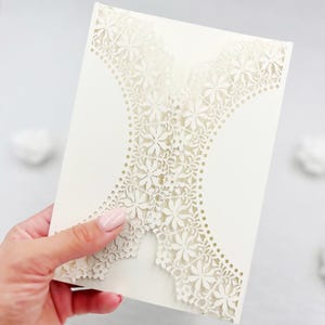 Peut inclure: Carte d'invitation ivoire avec des motifs floraux découpés au laser. La carte présente un motif ajouré de fleurs et de feuilles, créant un effet dentelle. La carte est tenue par une personne.