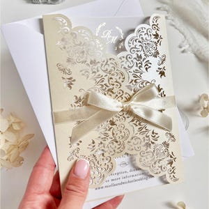 May include: Ivory wedding invitation with detailed laser-cut floral patterns and a satin ribbon bow. The invitation includes a matching envelope and features the initials "R M". The website address www.rosellaandmichaelwedding.com is visible.