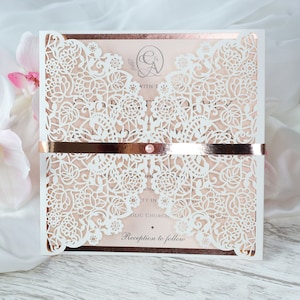 May include: White wedding invitation with a delicate floral lace design and a rose gold border. The invitation features a rose gold ribbon with a pearl embellishment and the text "Reception to follow".