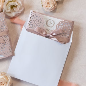 May include: A white envelope with a blush pink invitation inside. The invitation has a lace design and a blush pink ribbon with a bow. The text on the invitation reads "E & A" and "and A".