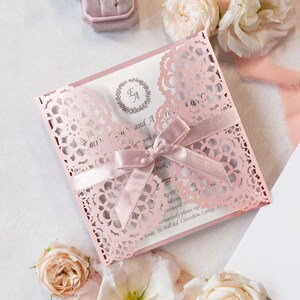 May include: A pink and white wedding invitation with a lace design and a pink ribbon. The invitation has the initials "E.A." in a wreath on the front. The text on the invitation reads "[Name] and [Name] with the date [Date] and the location [Location]. RSVP to [Email] or [Phone Number]."