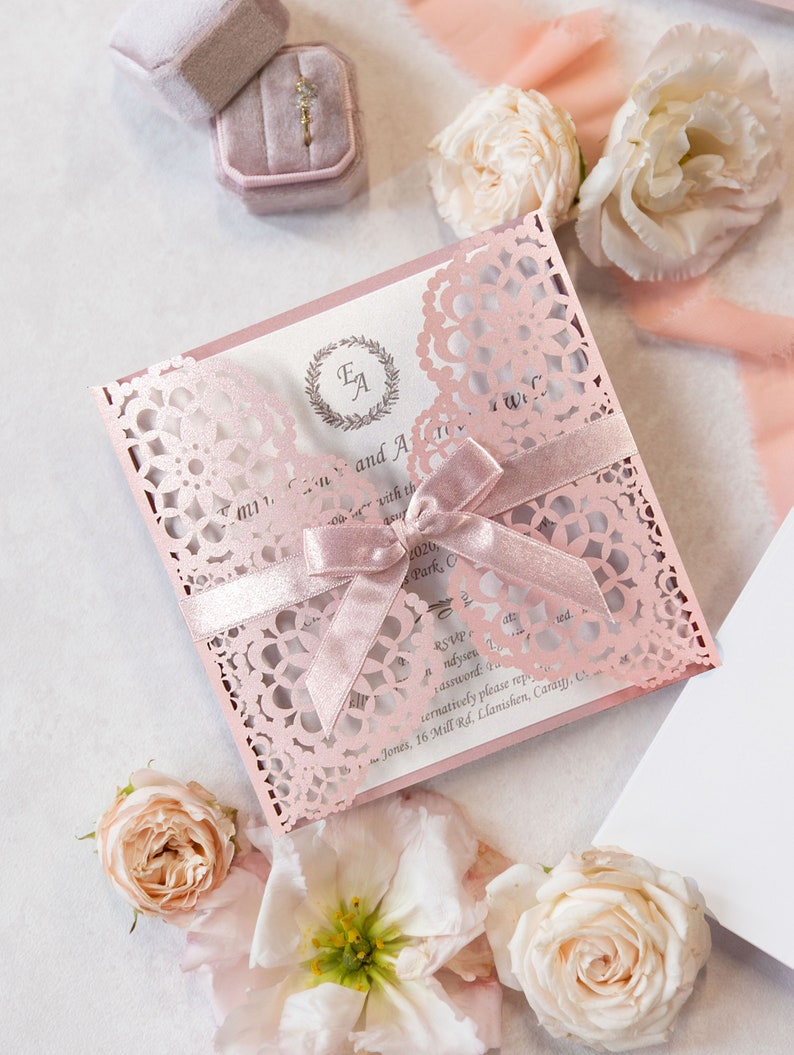 May include: A pink and white wedding invitation with a lace design and a pink ribbon. The invitation has the initials "E.A." in a wreath on the front. The text on the invitation reads "[Name] and [Name] with the date [Date] and the location [Location]. RSVP to [Email] or [Phone Number]."