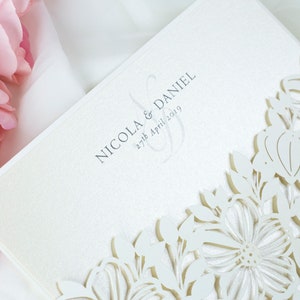 May include: Cream-coloured wedding invitation with a laser-cut floral pattern. The text reads "NICOLA & DANIEL" and "27th April 2019". The design incorporates delicate cut-out flowers. Pink flowers are visible in the background, adding a soft touch.