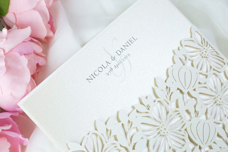 May include: Cream-coloured wedding invitation with a laser-cut floral pattern. The text reads "NICOLA & DANIEL" and "27th April 2019". The design incorporates delicate cut-out flowers. Pink flowers are visible in the background, adding a soft touch.