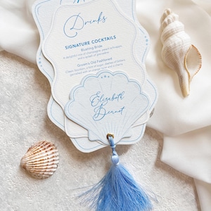 May include: A die-cut menu card with light blue ink text reading Menu, Drinks, Signature Cocktails, Blushing Bride, Groom's Old Fashioned, and Elizabeth Bennet. The card is shaped like a shell, features a gold eyelet, and a light blue tassel.