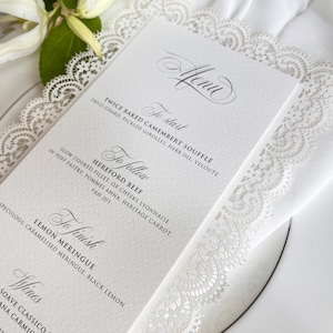 Può includere: Un menu in carta bianca con testo nero che presenta le sezioni Menu, To start, To follow, To finish e Wines. Il menu è incorniciato da un intricato bordo in pizzo bianco e poggia su un piatto bianco, decorato con fiori bianchi.